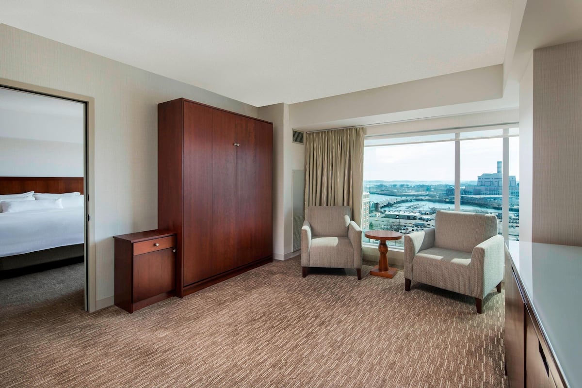 A hotel room with a bed, a wardrobe, two armchairs, a small table, and a large window with a city view.