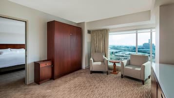 A hotel room with a bed, a wardrobe, two armchairs, a small table, and a large window with a city view.