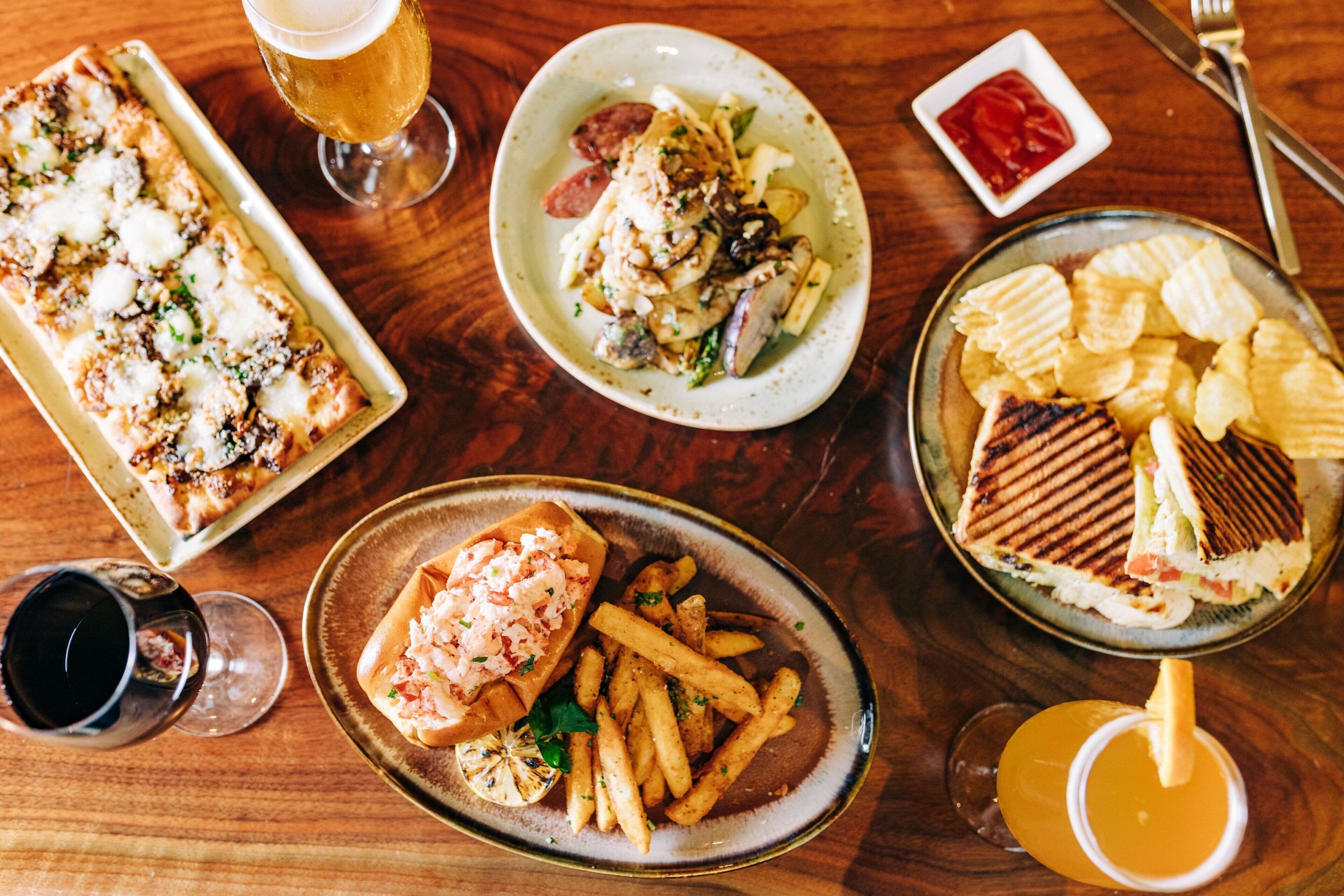 Variedad de platos como pizza, papas fritas y un sándwich a la plancha, junto con bebidas sobre una mesa de madera.
