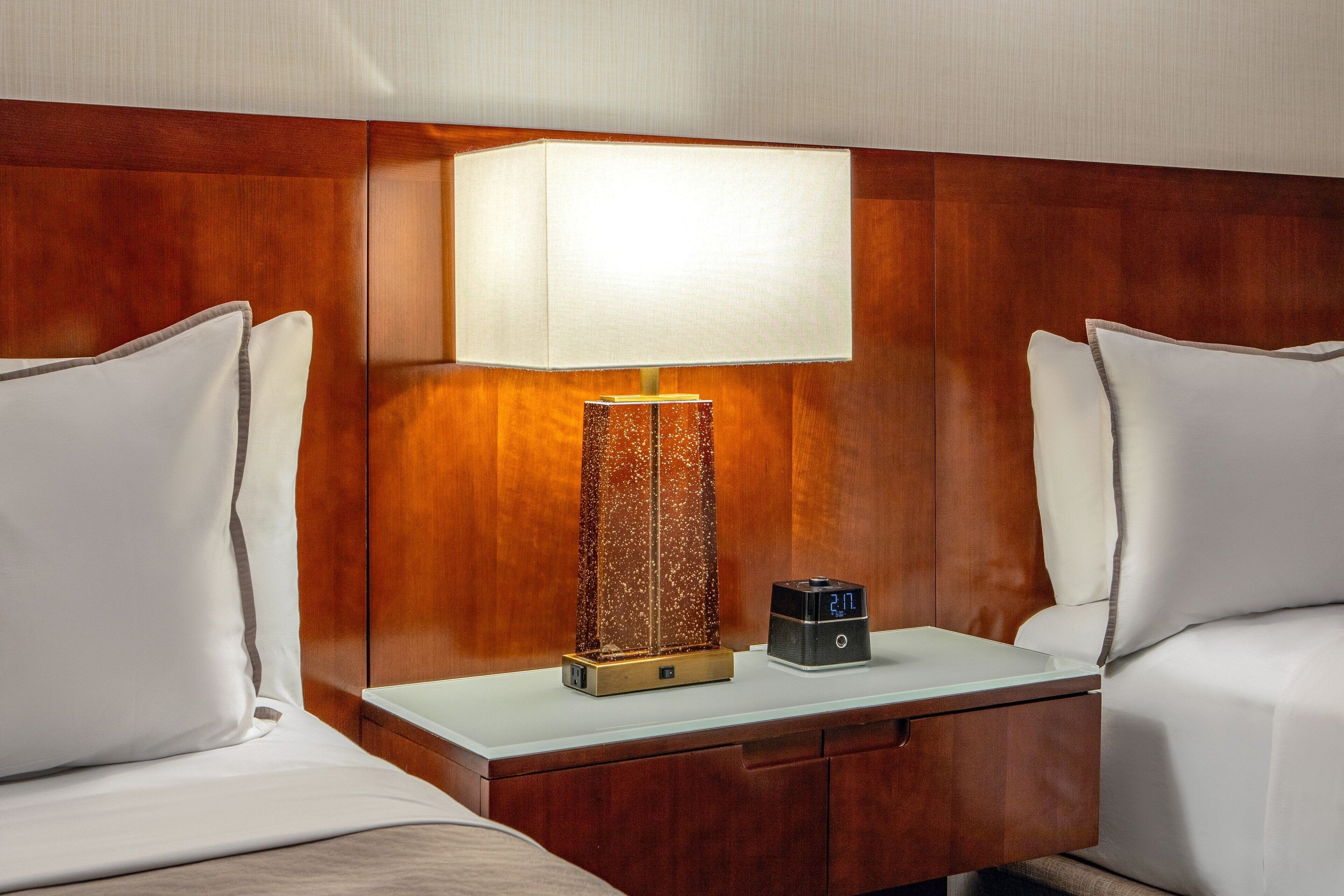 A hotel room with a wooden headboard, a bedside table with a lamp, a digital clock, and two white pillows.