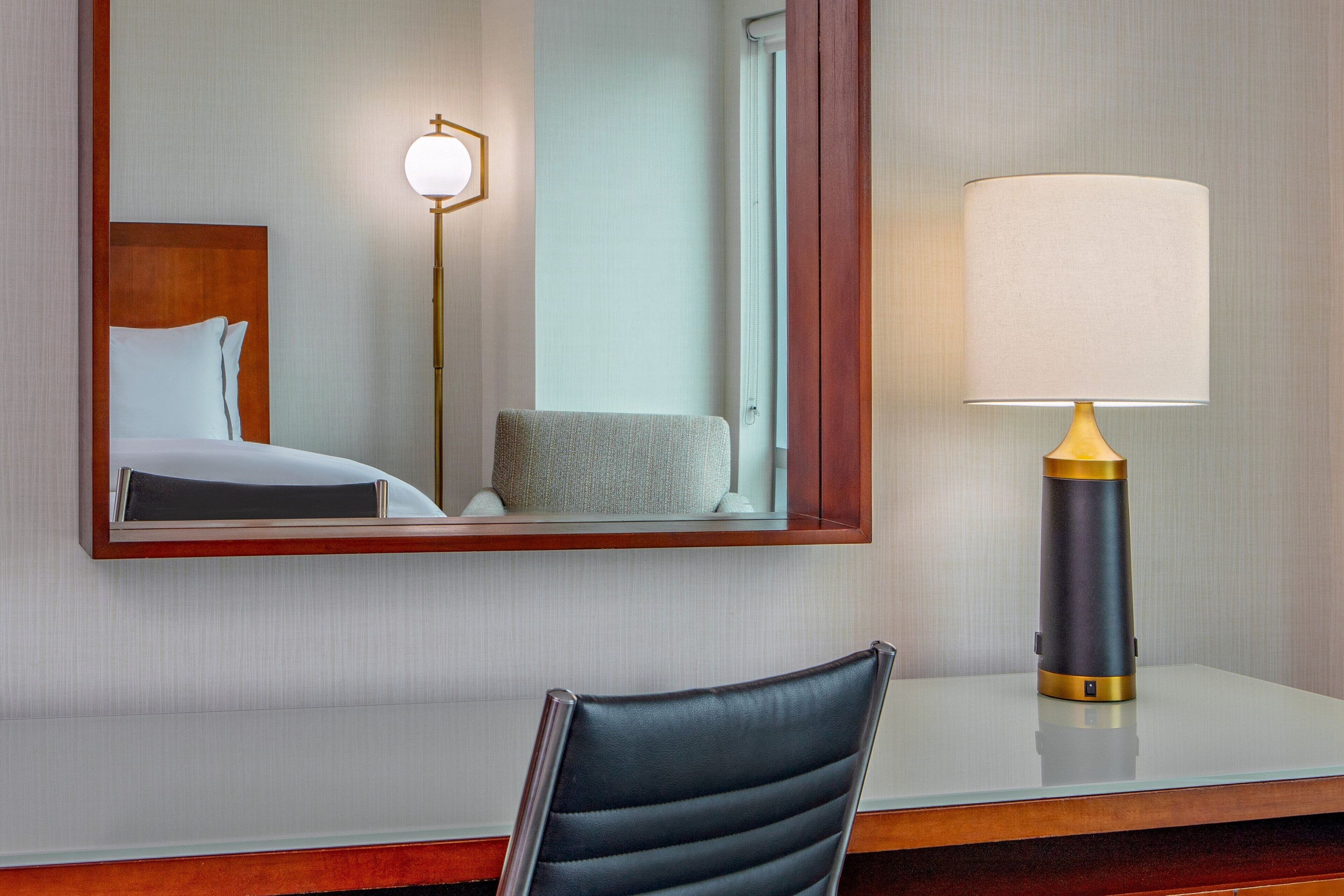 A hotel room with a bed, a chair, a desk, a lamp, and a mirror.