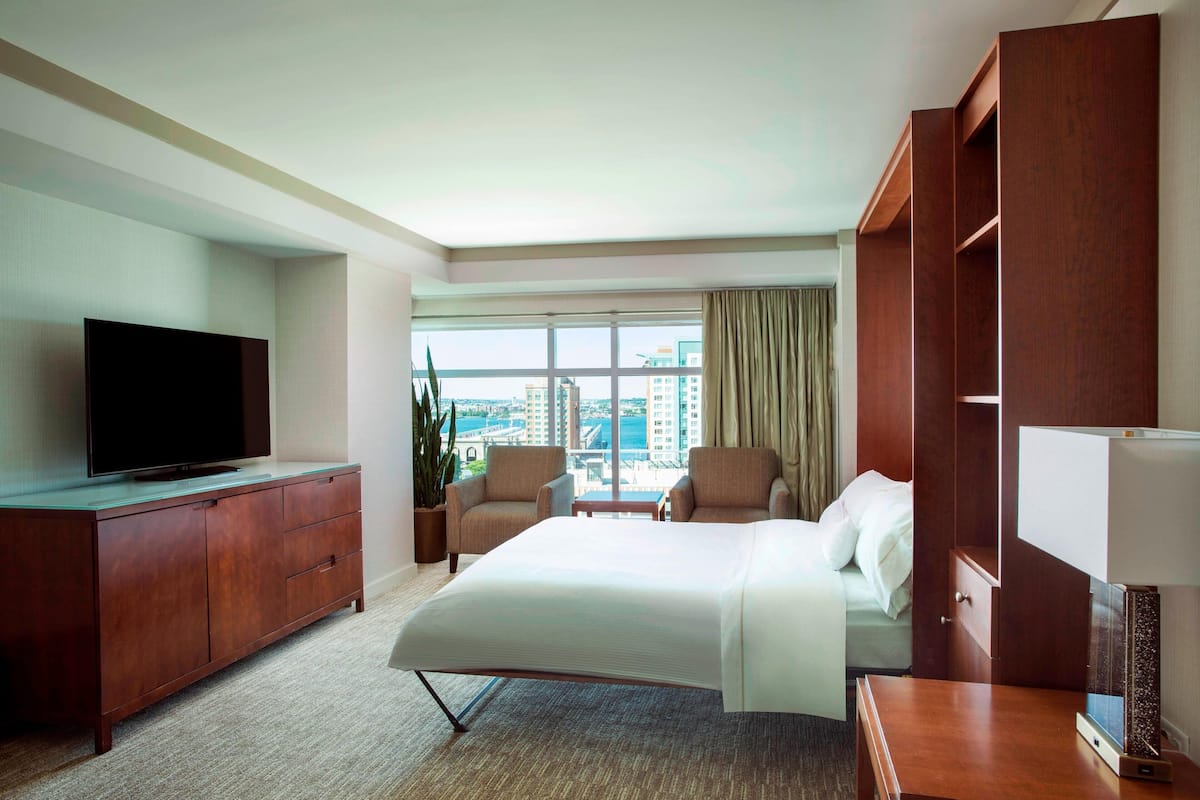 A hotel room with a large bed, a TV on a wooden cabinet, two armchairs, a desk, and a view of the city through large windows.