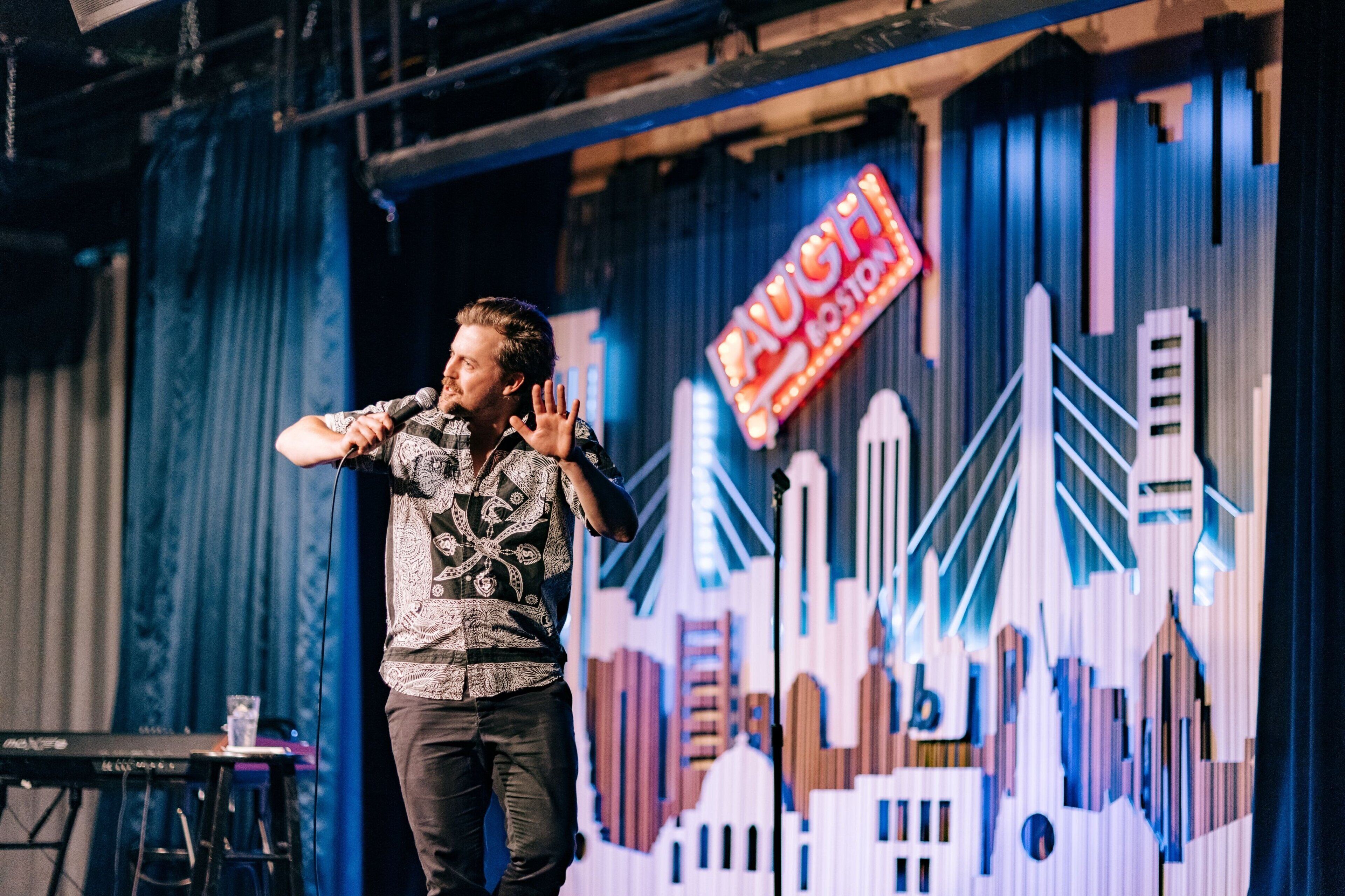 A person on stage with a microphone, a keyboard, and a cityscape backdrop.