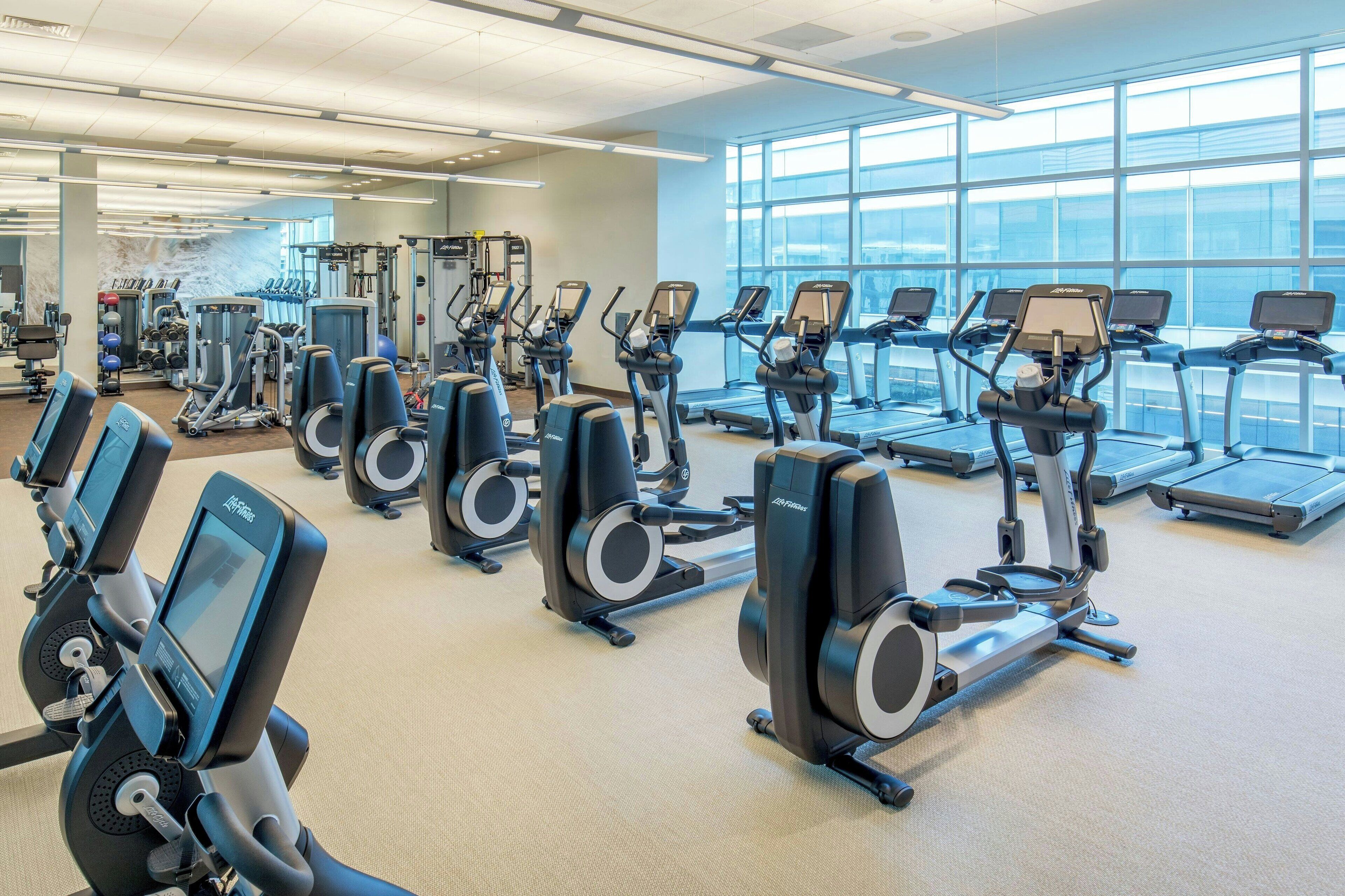 Et fitnesscenter med flere løbebånd og crosstrainere, store vinduer og forskellig træningsudstyr.