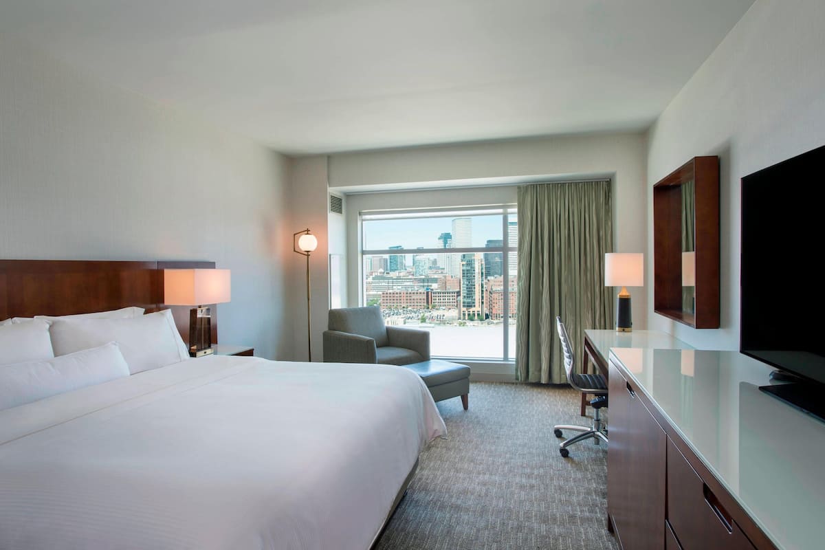 A hotel room with a large bed, a desk with a chair, a TV, and a view of the city through a large window.
