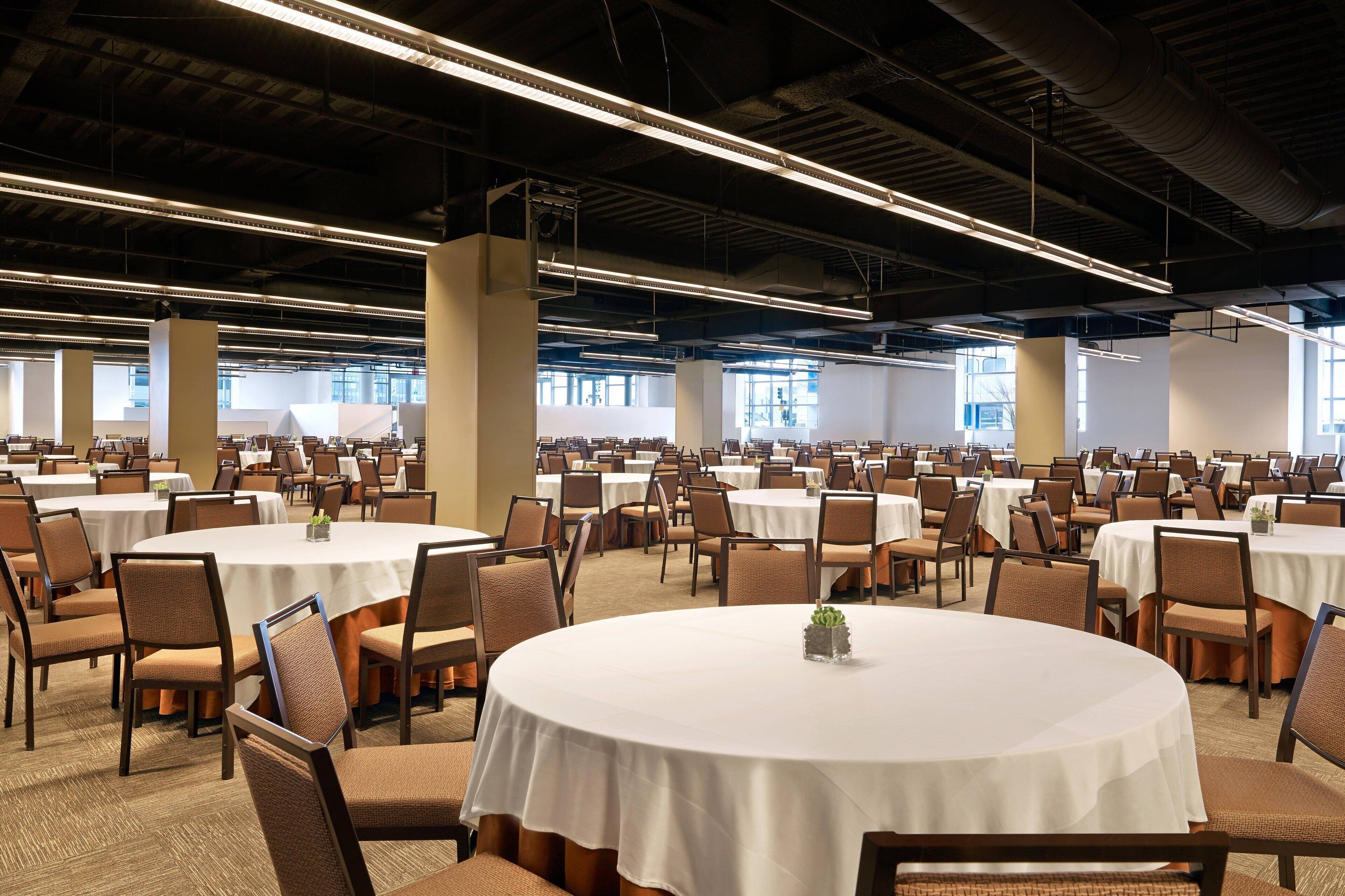 A large conference room with round tables and chairs, high ceilings with exposed beams, and large windows allowing natural light.
