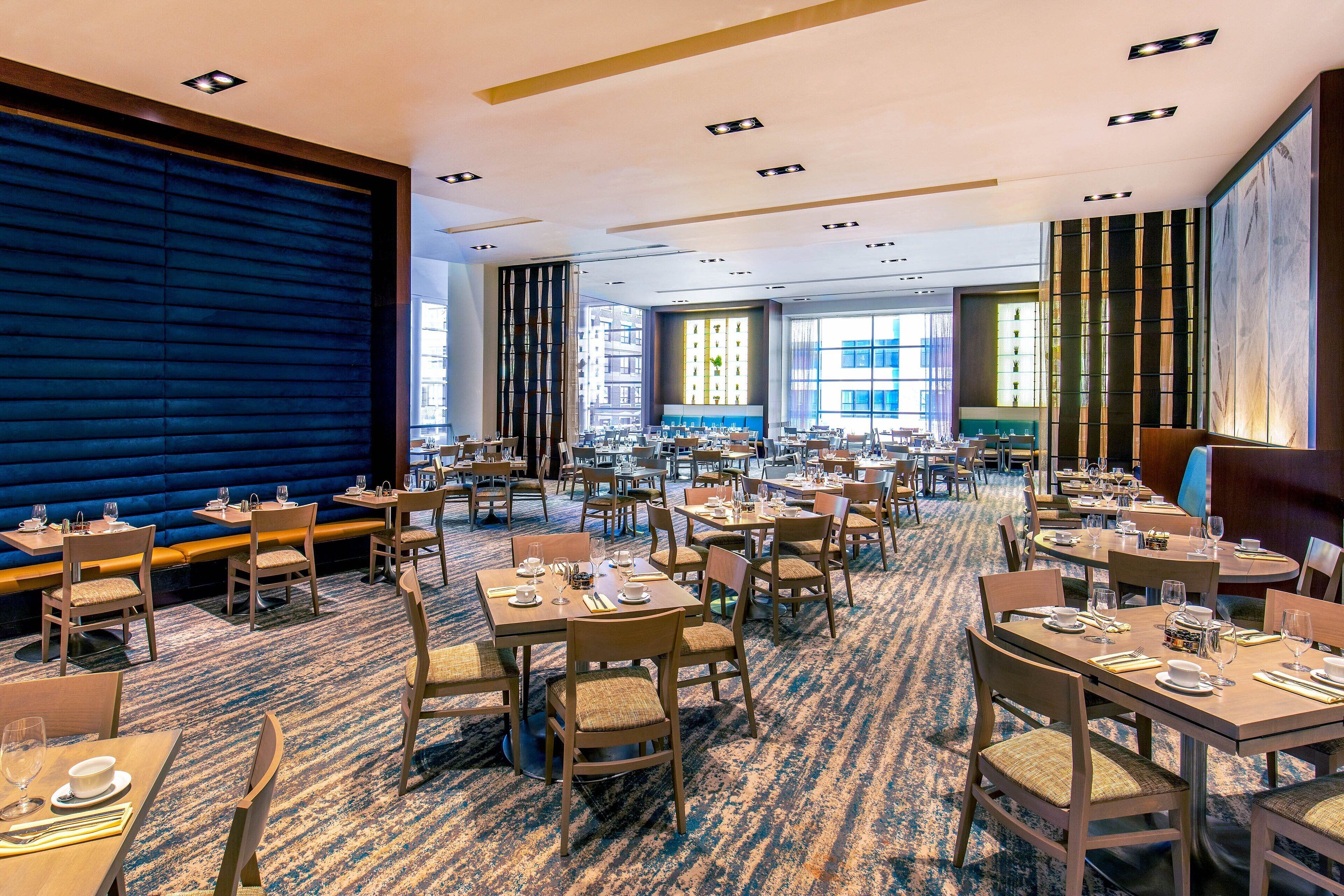 Een ruime hotel eetruimte met meerdere tafels en stoelen, grote ramen en een blauwe accentmuur.