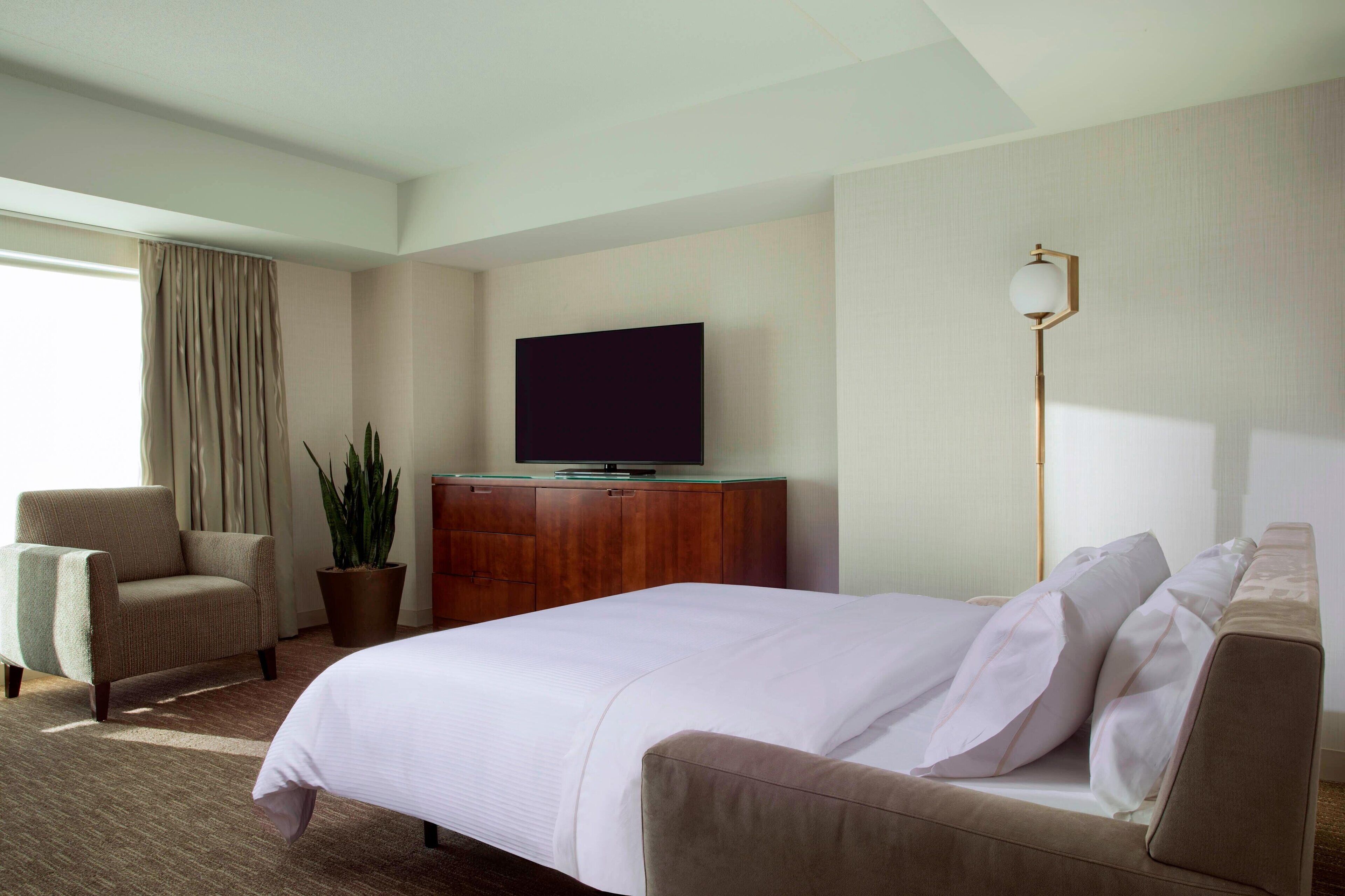A hotel room with a large bed, a television on a wooden cabinet, a potted plant, a chair, and a floor lamp.