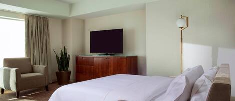 A hotel room with a large bed, a television on a wooden cabinet, a potted plant, a chair, and a floor lamp.