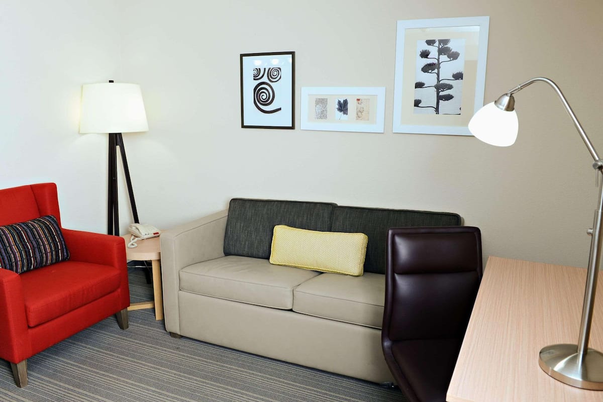 A hotel room with a red armchair, a beige sofa, a small table, a lamp, and framed artwork on the wall.