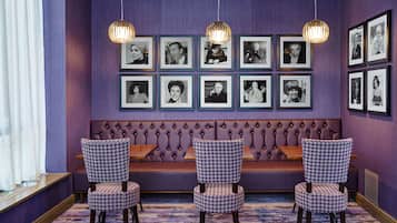 A room with a purple wall, a tufted bench, and framed black and white photographs. There are three patterned chairs and two hanging pendant lights.
