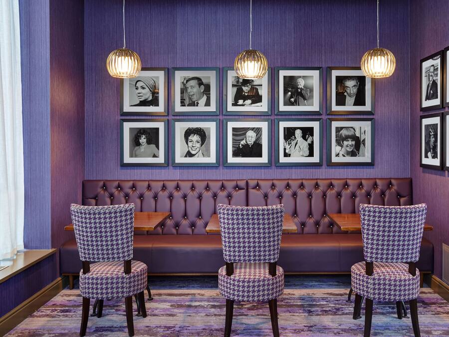 A room with a purple wall, a tufted bench, and framed black and white photographs. There are three patterned chairs and two hanging pendant lights.