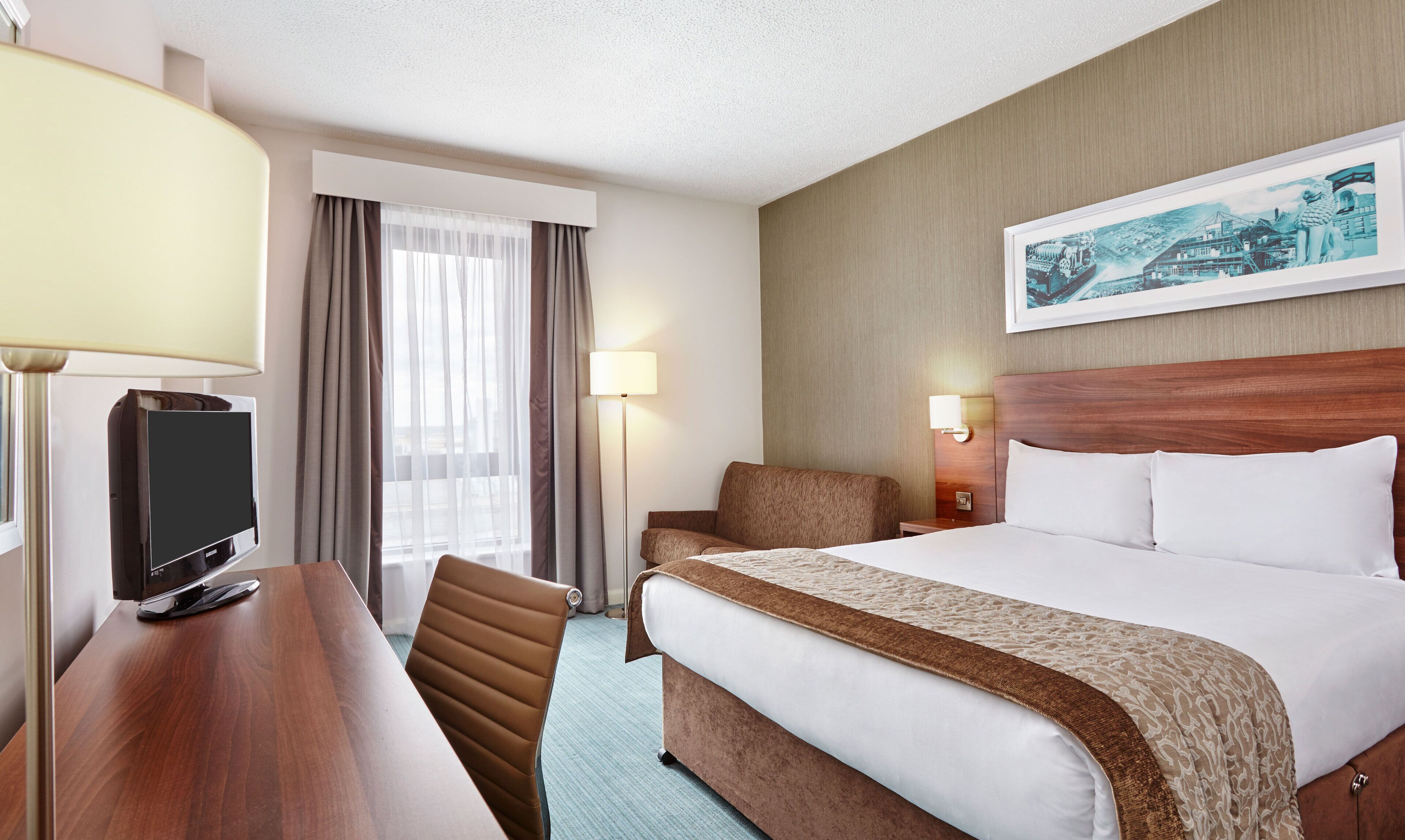 A hotel room with a bed, a desk with a TV, a chair, a lamp, and a painting on the wall.