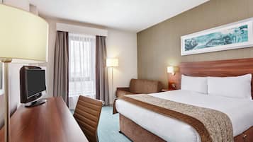 A hotel room with a bed, a desk with a TV, a chair, a lamp, and a painting on the wall.