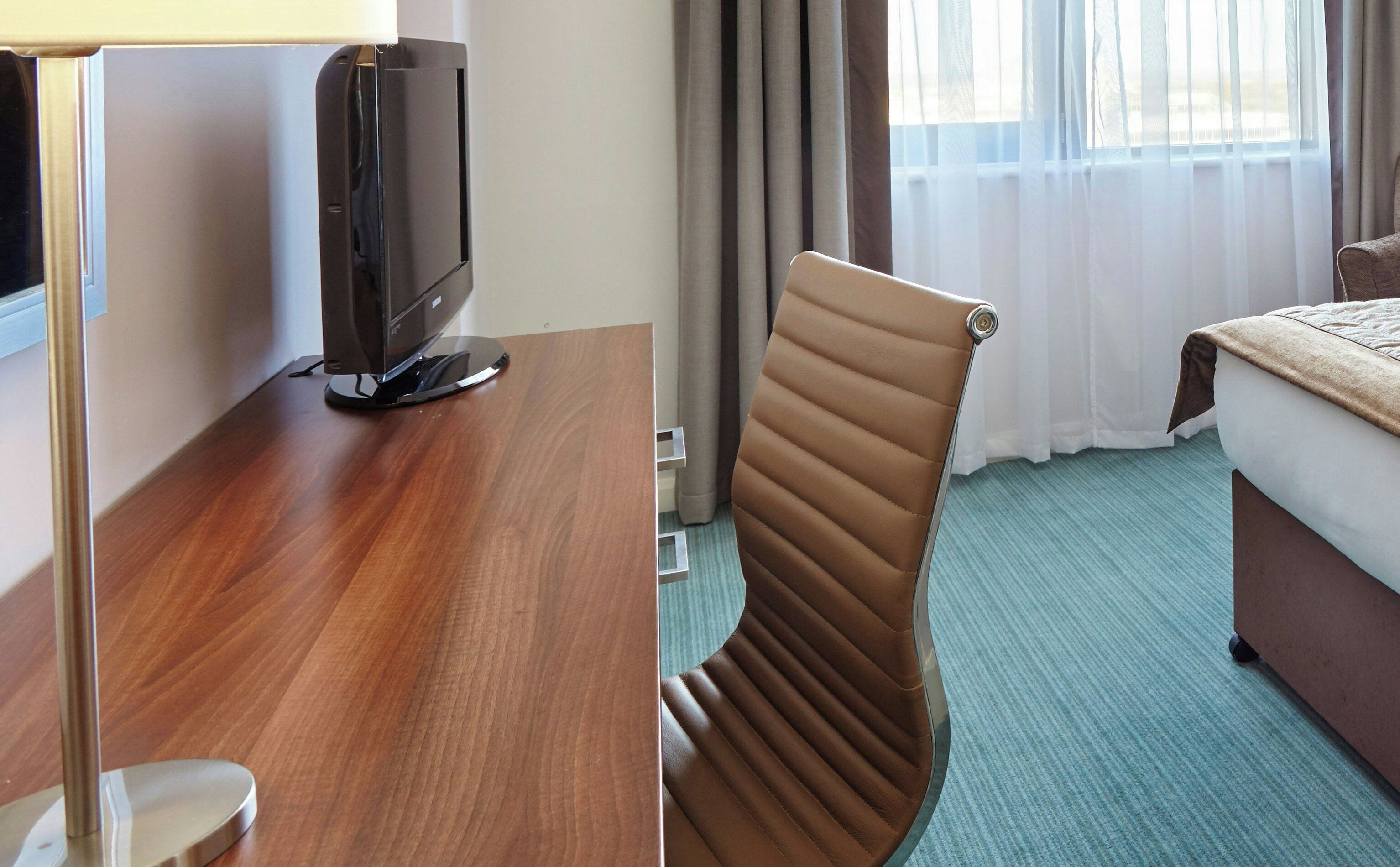 A hotel room with a wooden desk, a television, a brown leather chair, a bed, and a window with curtains.