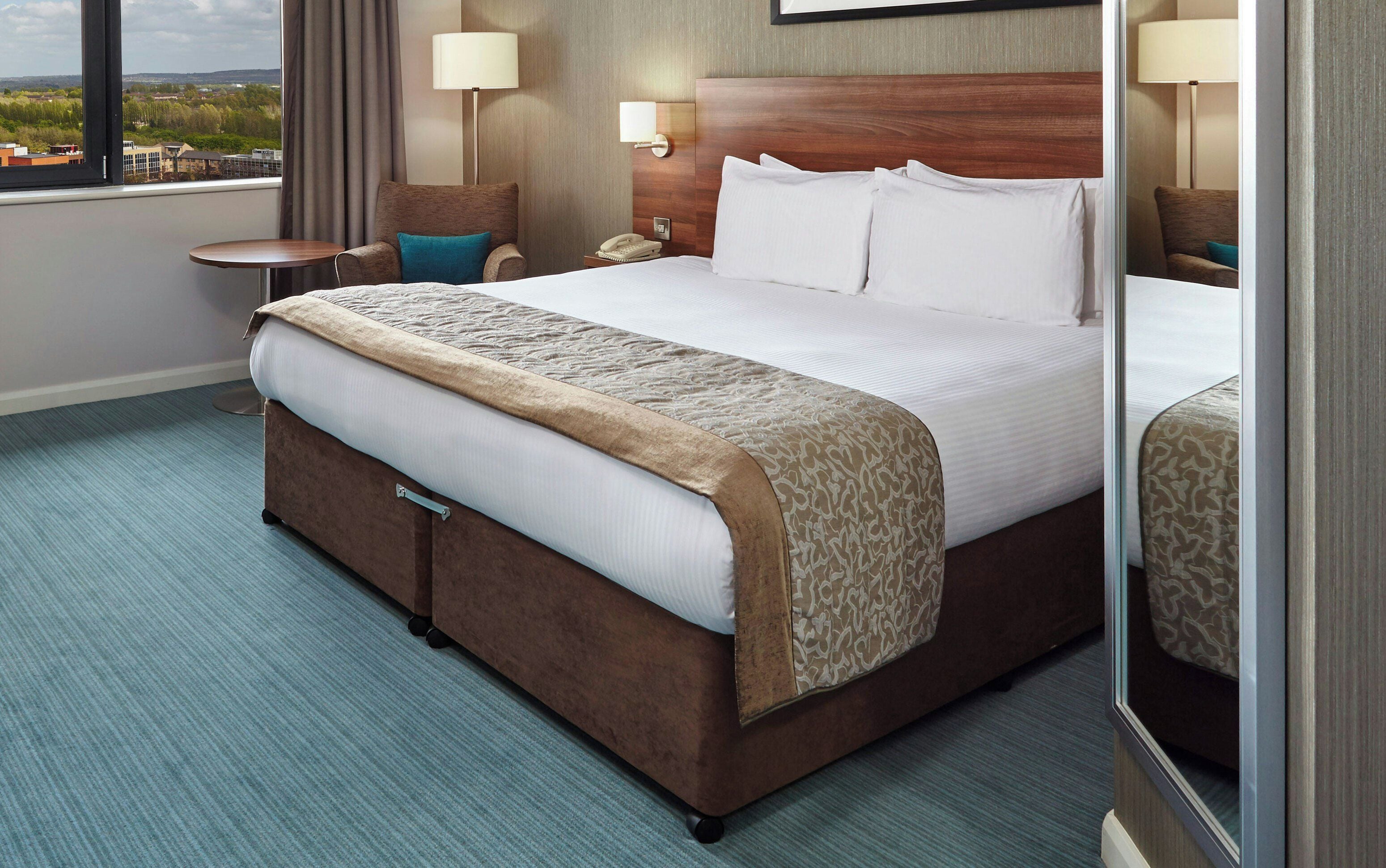 A hotel room with a large bed, a bedside table, a lamp, and a view of the cityscape.
