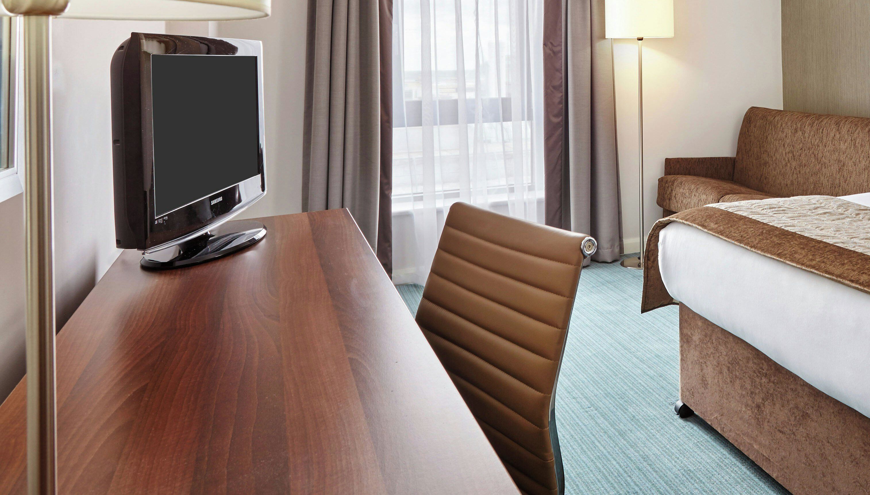 A hotel room with a wooden desk, a television, a brown leather chair, a bed with a brown headboard, and a window with curtains.