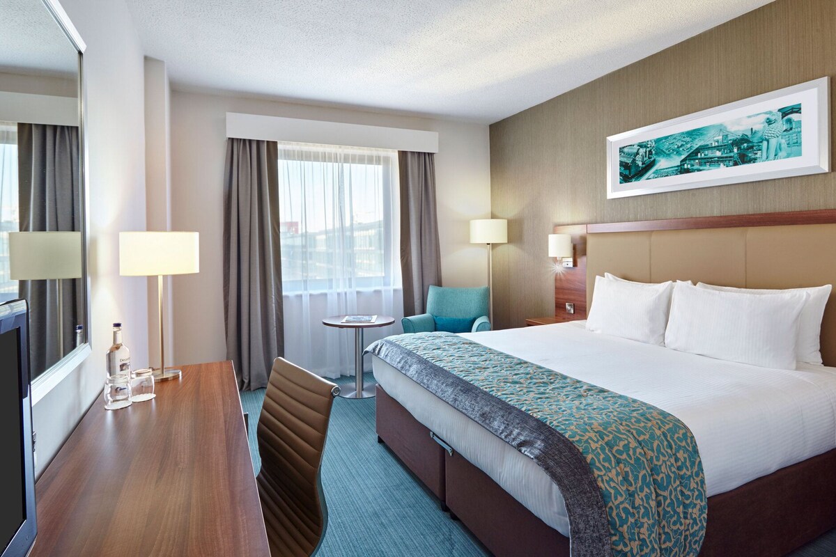 A hotel room with a bed, a desk, a chair, a small table, and a window with curtains.