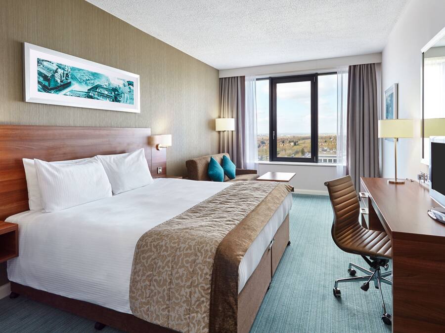 A hotel room with a large bed, a desk with a computer, a chair, a TV, and a window with a view.