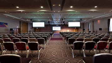 A conference room with rows of chairs, a stage, a projector screen, and a carpeted floor.