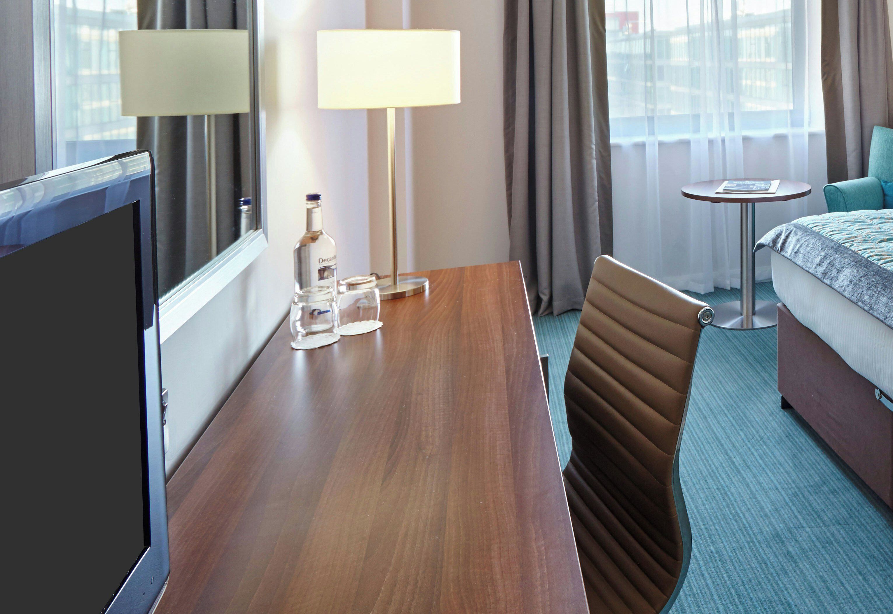 Una habitación de hotel con un escritorio de madera, una silla, una cama, una mesita y una lámpara.