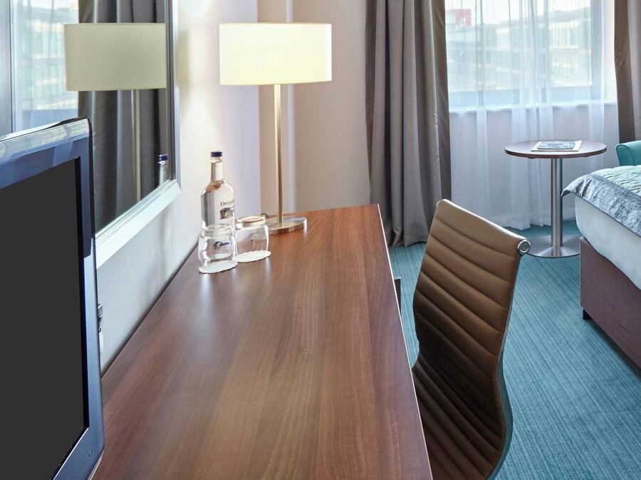 Una habitación de hotel con un escritorio de madera, una silla, una cama, una mesita y una lámpara.
