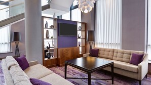 A modern living room with a sofa, armchair, and a coffee table. There is a TV mounted on the wall, a bookshelf, and a large window with curtains.
