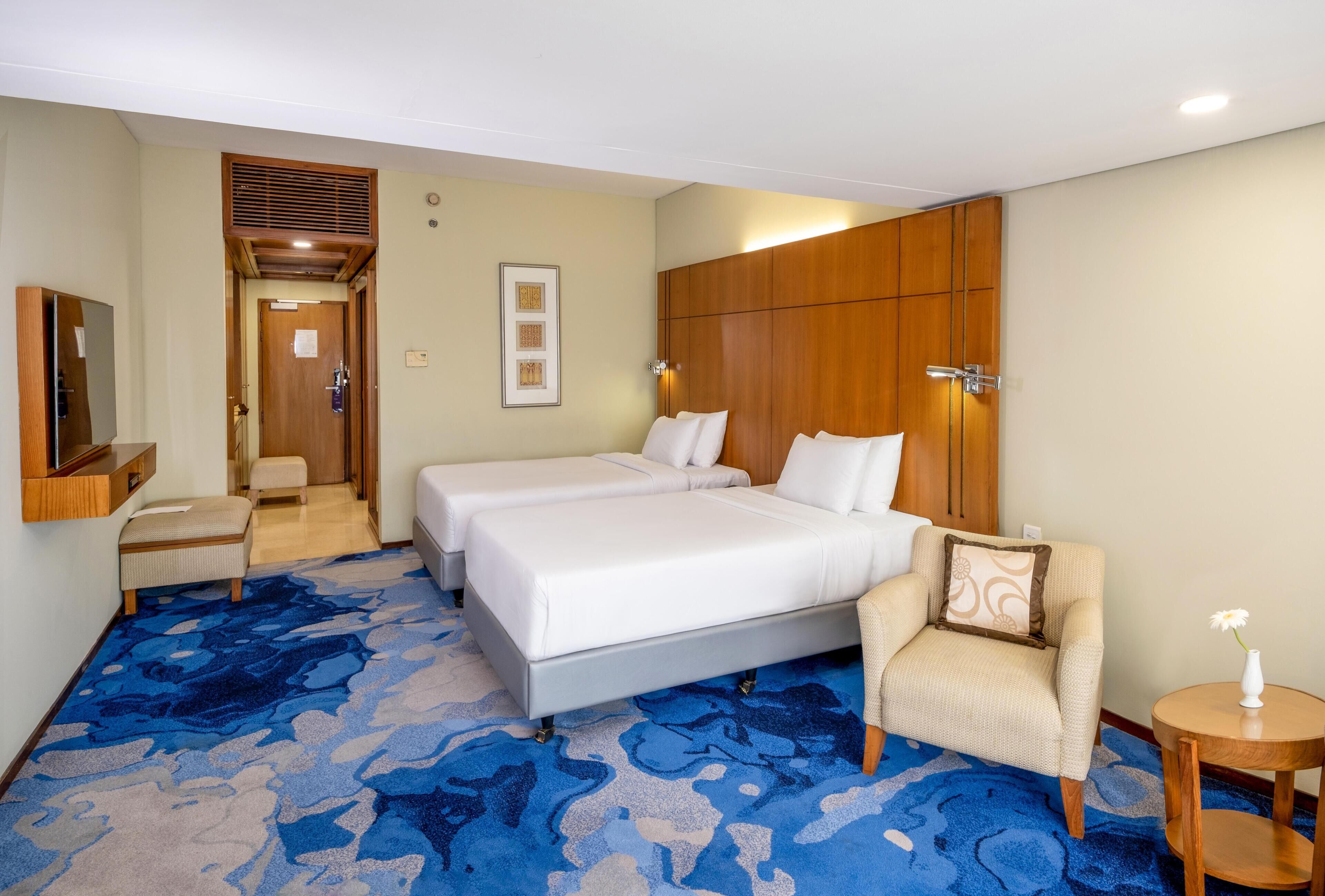 A hotel room with a large bed, a chair, a small table, a TV, and a wooden wardrobe.