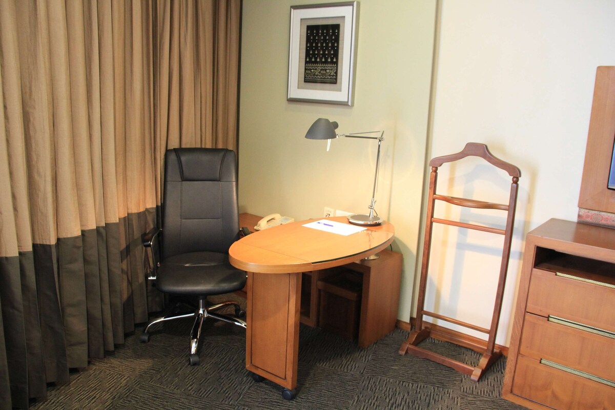 Quarto de hotel com uma escrivaninha, uma cadeira de escritório preta, uma luminária, um telefone, uma cadeira de madeira e um pequeno armário.