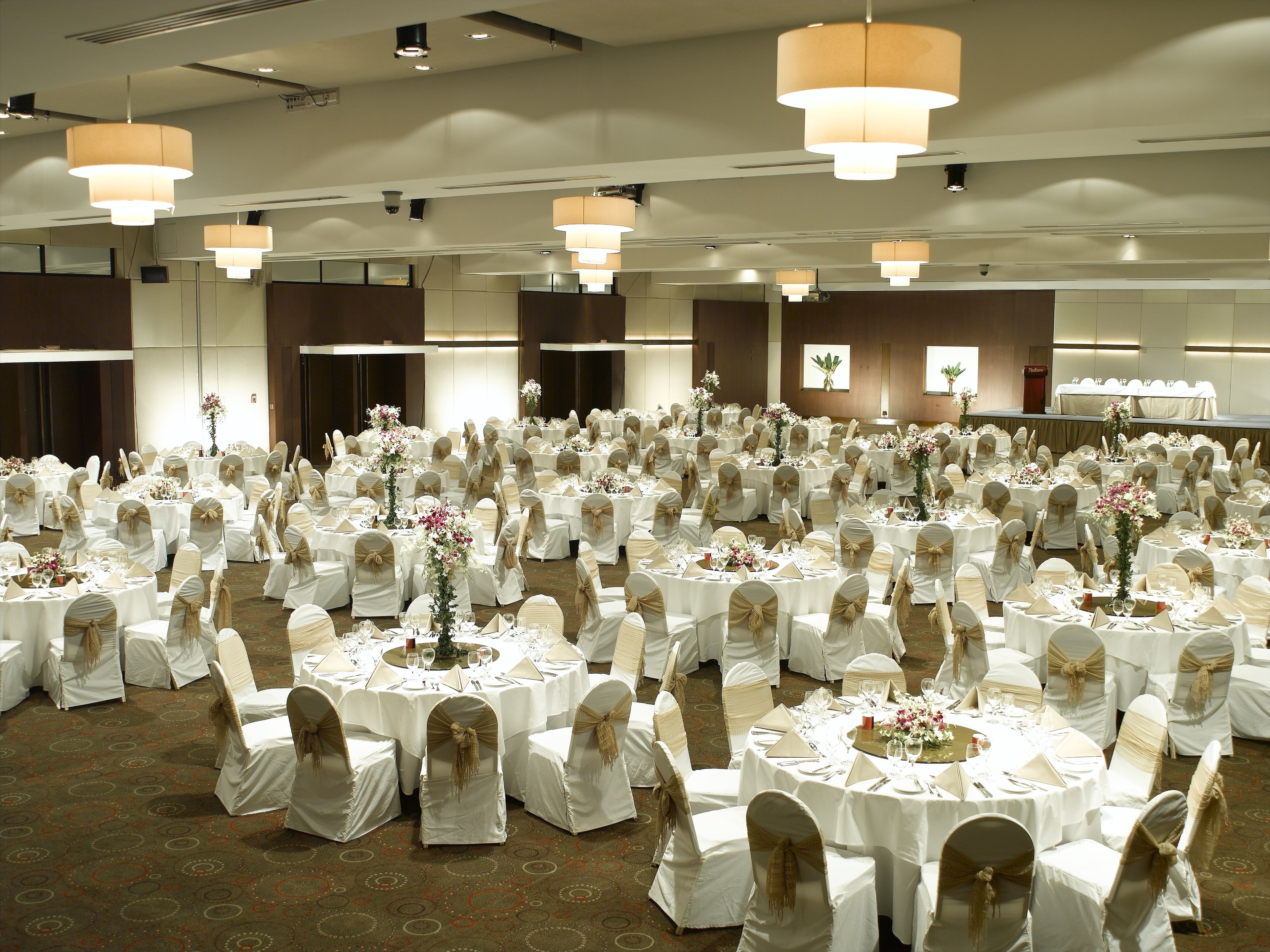 Une vaste salle de banquet, avec des tables rondes dressées pour un événement formel, agrémentée de nappes blanches et de chaises ornées de rubans dorés.