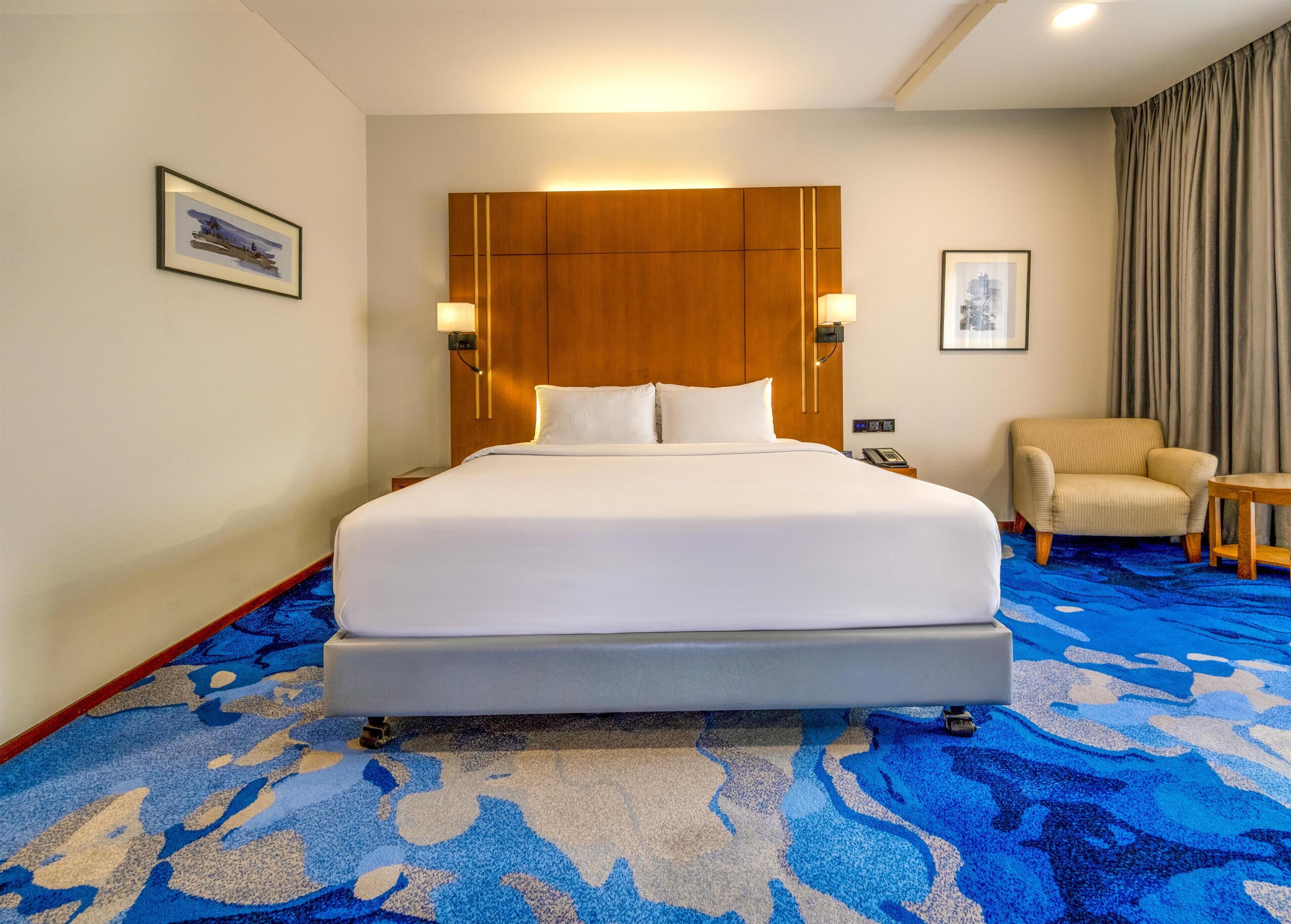 Une chambre d’hôtel avec un grand lit, une tête de lit en bois, deux lampes fixées au mur, une chaise et une moquette à motifs bleus et blancs.