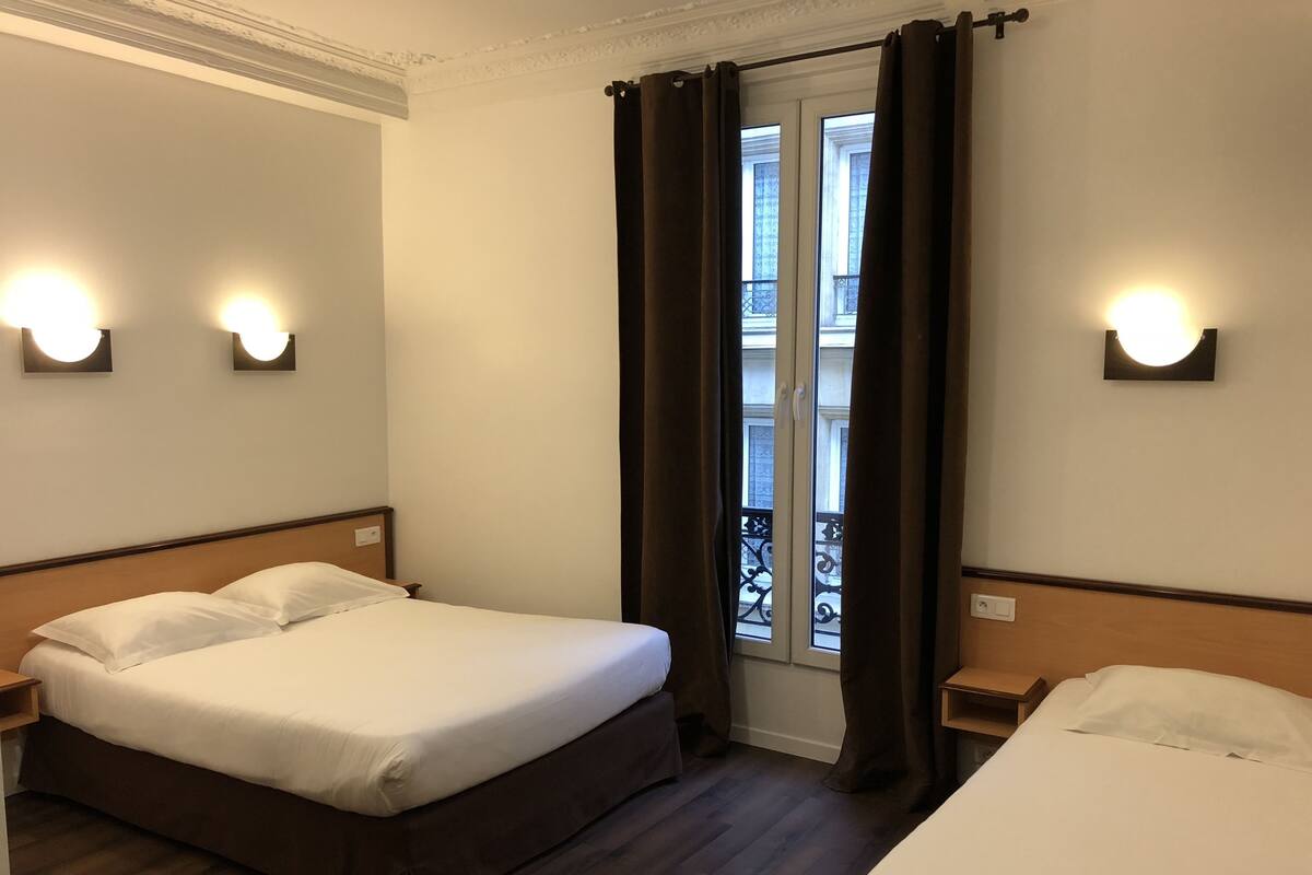 A hotel room with a double bed, a smaller bed, two bedside tables, and a window with curtains.