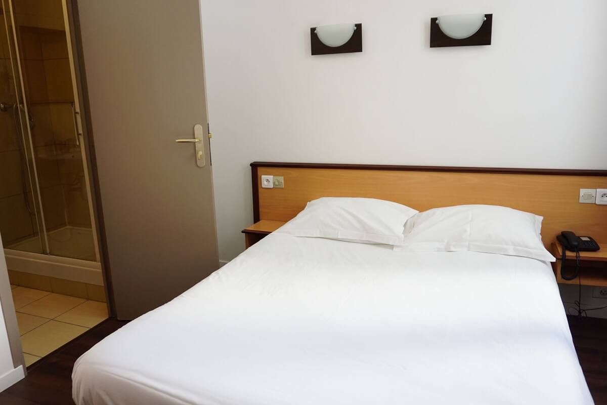 A hotel room with a bed, a bedside table with a phone, and a wall-mounted light fixture.
