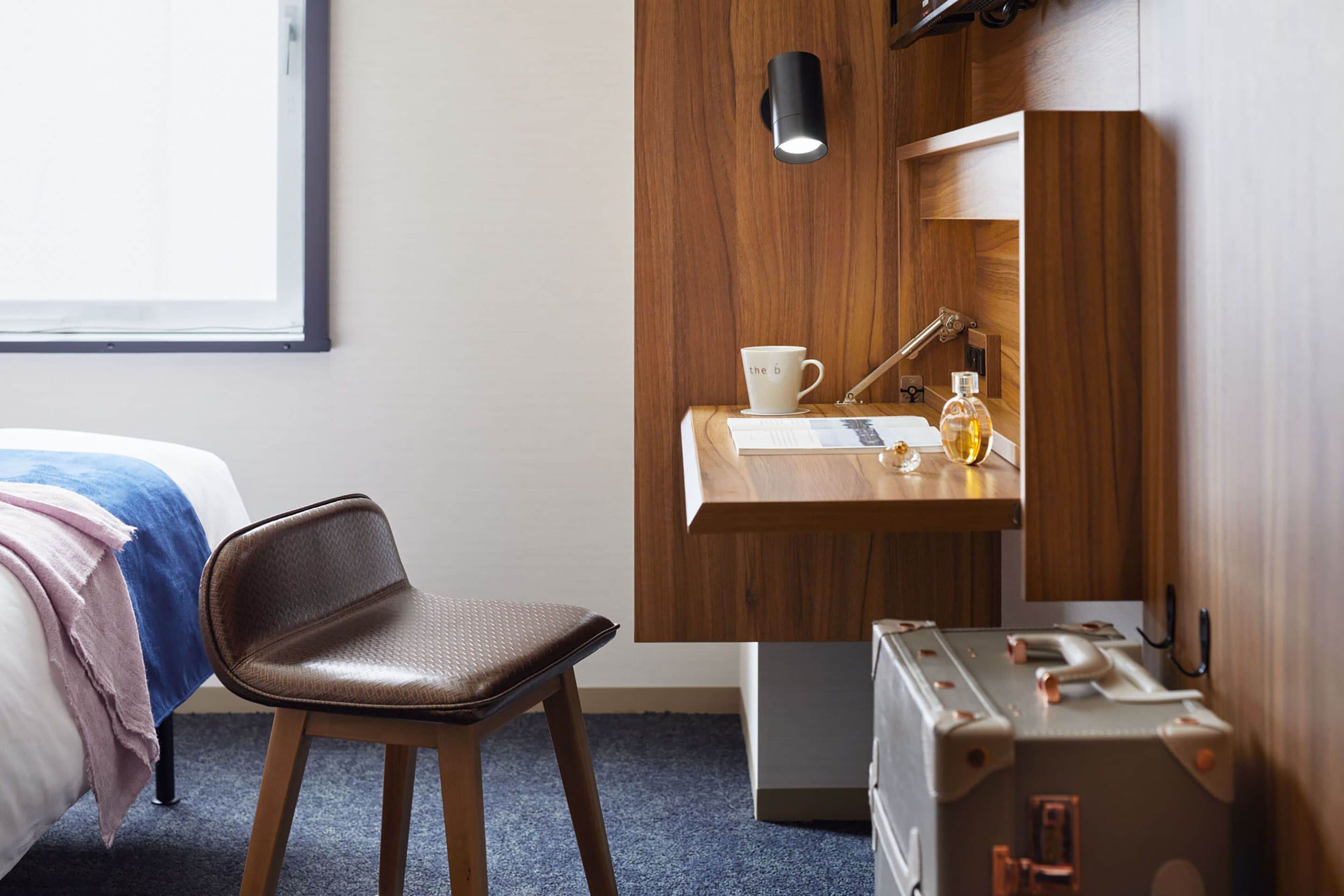 A hotel room with a bed, a chair, a desk with a cup, a lamp, and a suitcase.