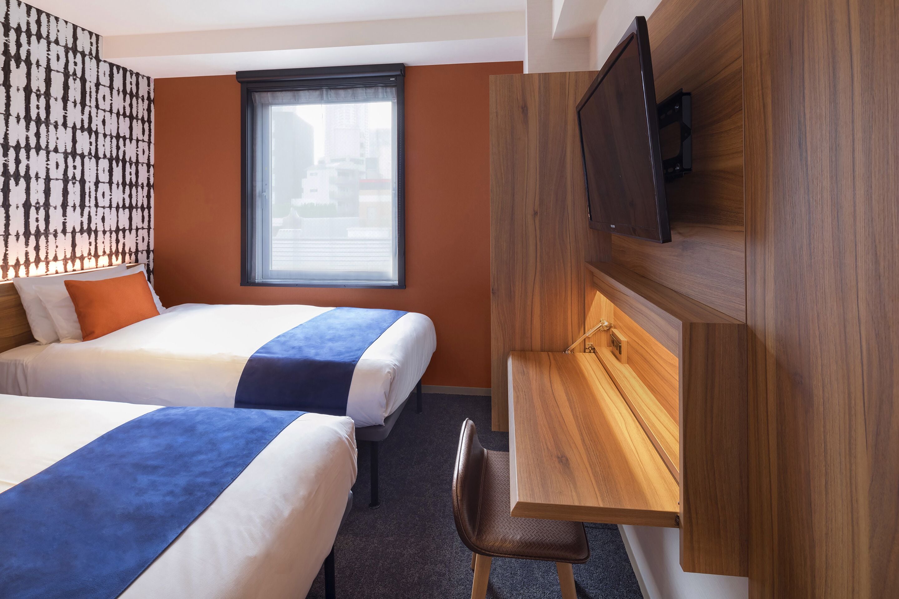 A hotel room with two beds, a desk, a chair, a TV mounted on a wooden panel, and a window with blinds.