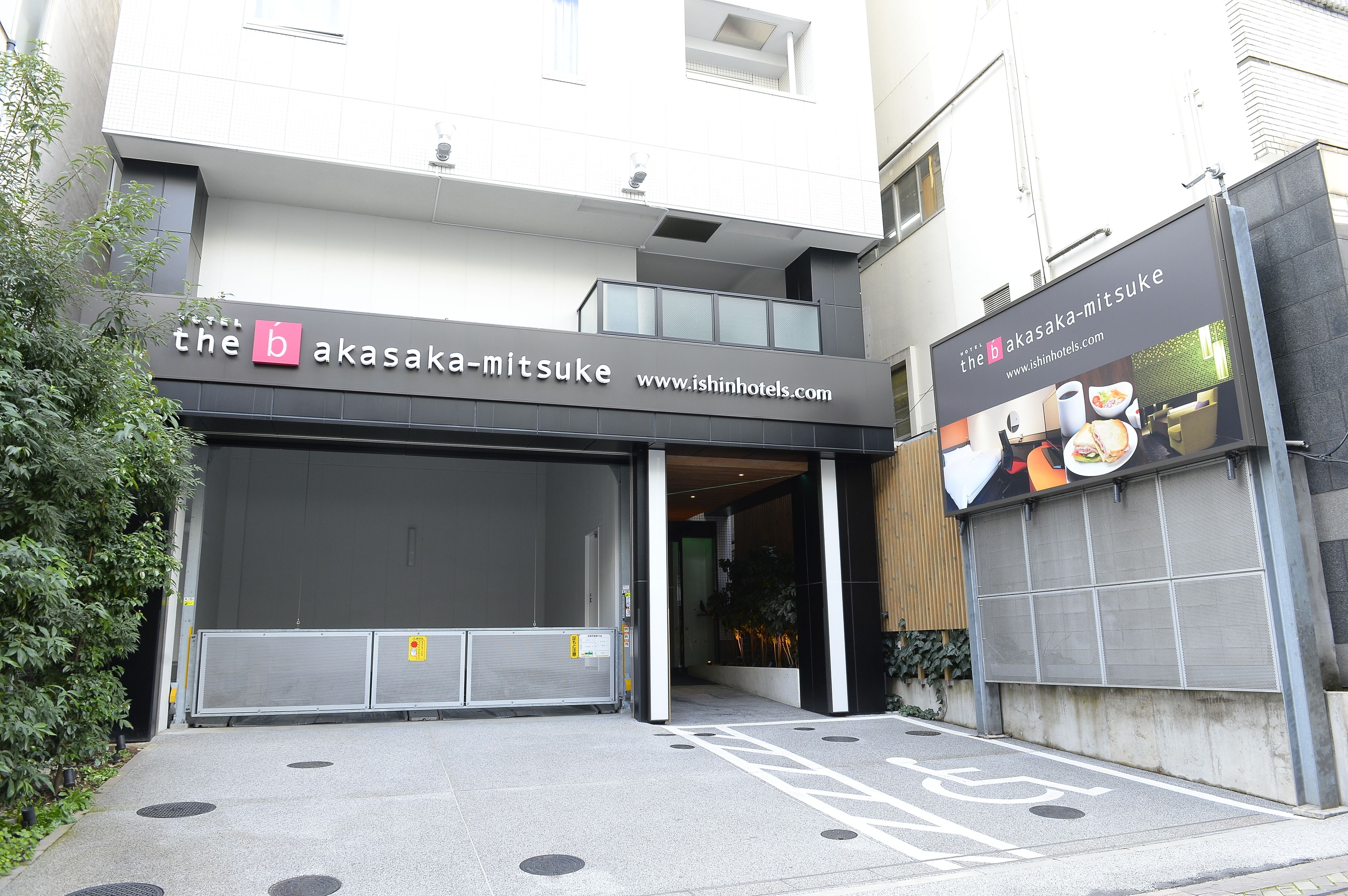 A hotel entrance with a closed garage door, a sign for 'The b akasaka-mitsuke', and a website URL.