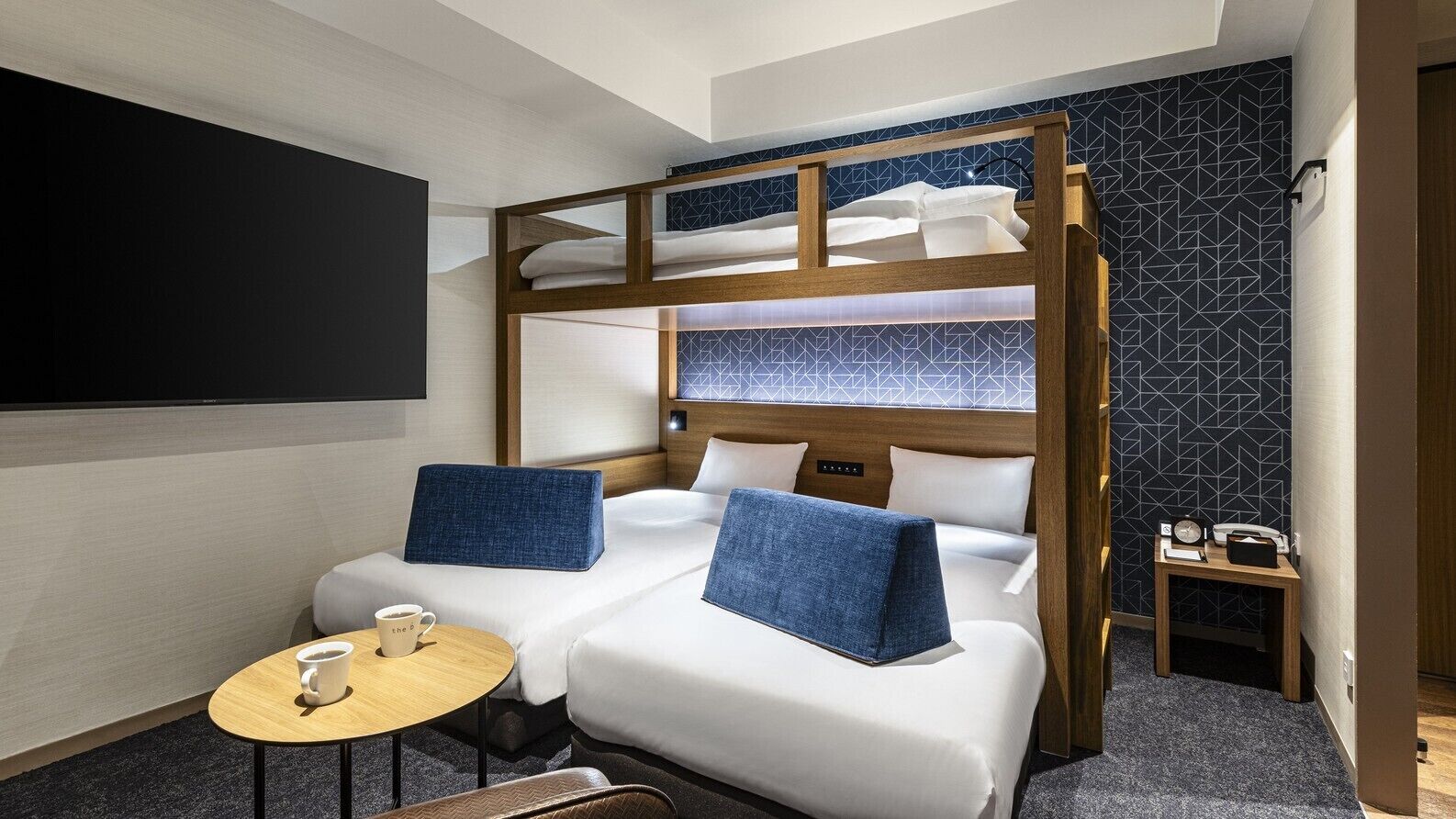 A hotel room with a bunk bed, two single beds, a small table with a cup, a bedside table with a phone, and a wall-mounted TV.