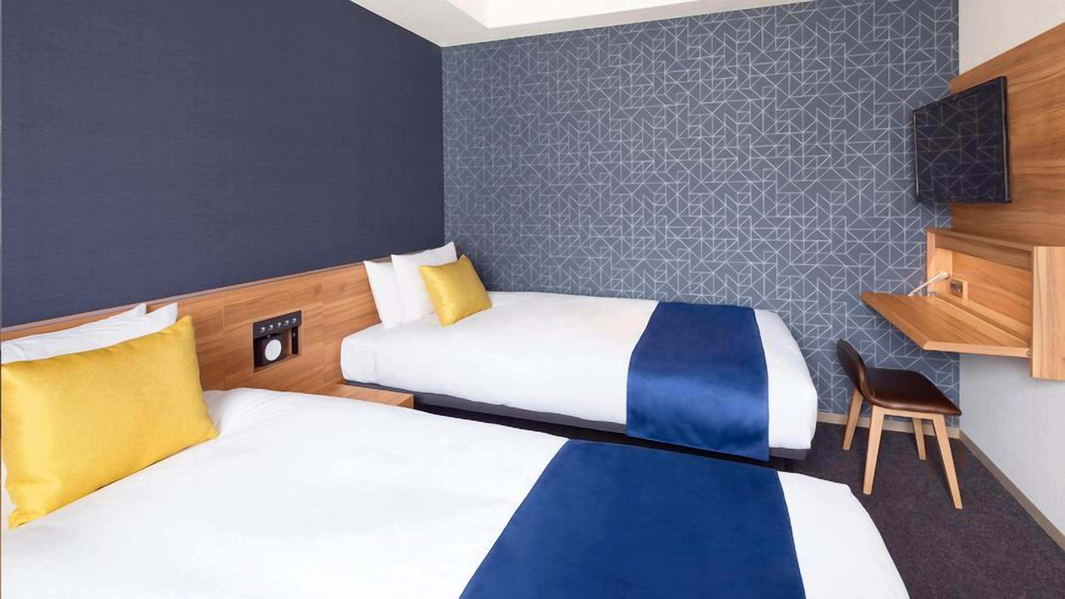 A hotel room with two beds, a desk, a chair, a TV, and a wall clock.