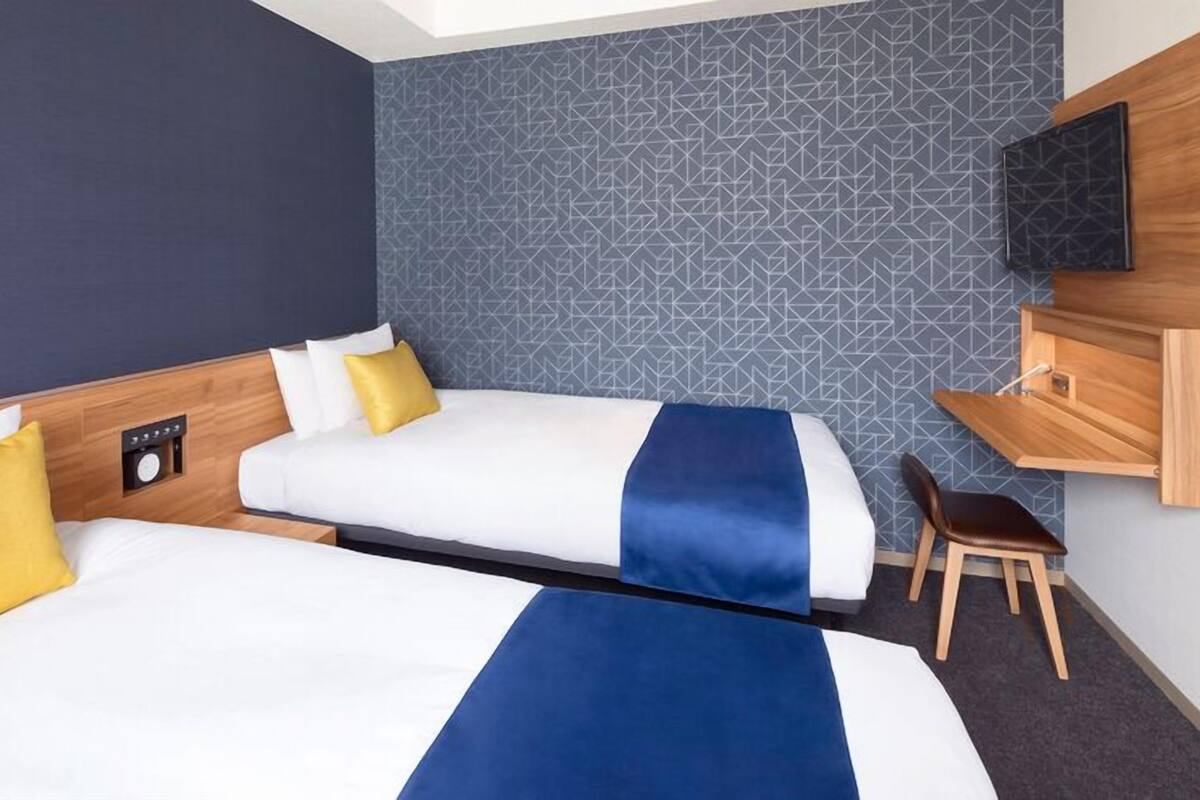 A hotel room with two beds, a desk, a chair, a TV, and a wall clock.