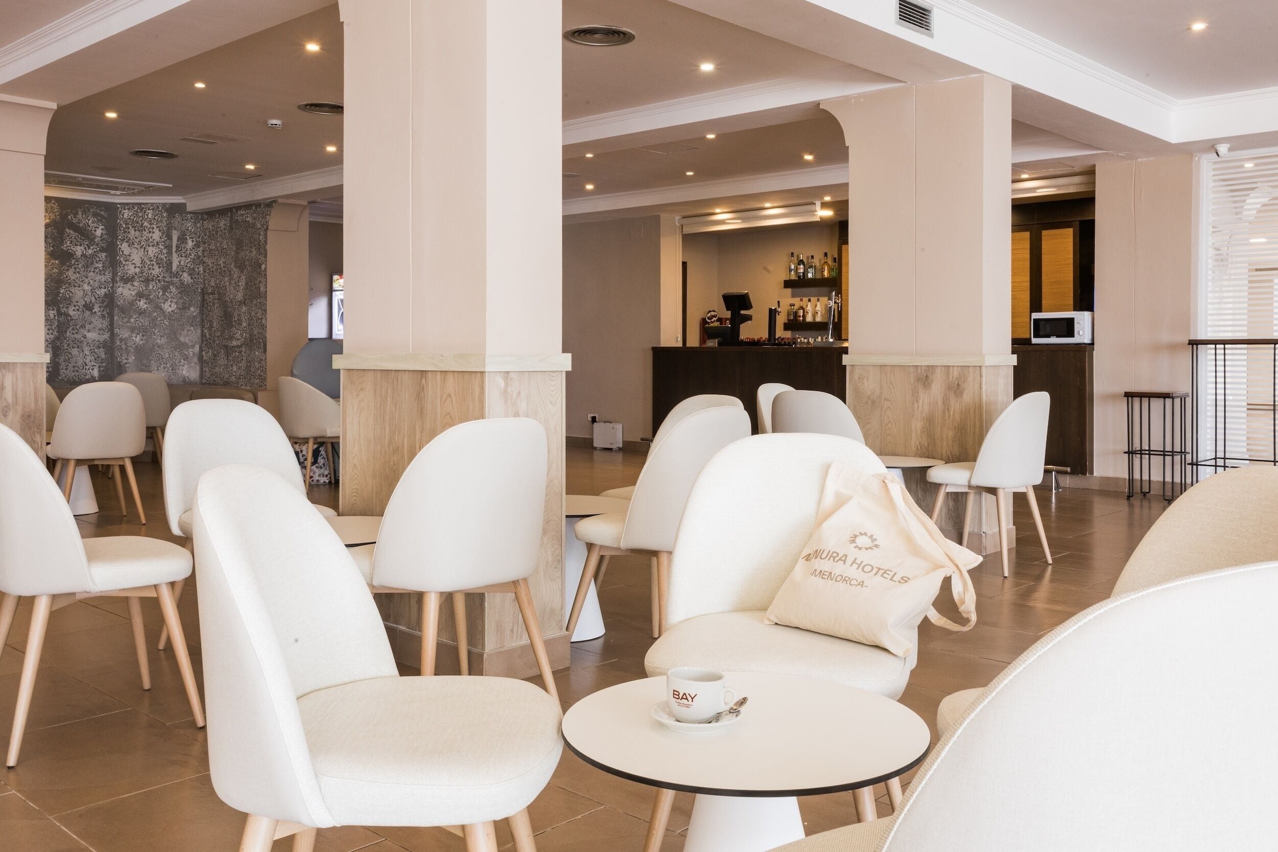 A modern hotel lobby with white chairs and tables, a reception desk, and a bar area.