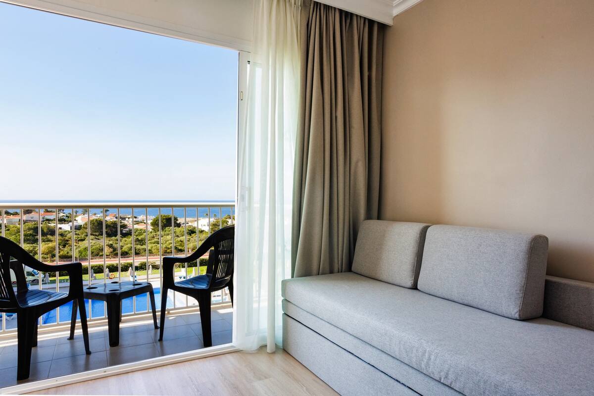 A balcony with a sofa and two chairs, overlooking a pool and a view of the sea.