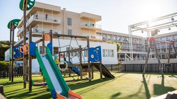 Un parque infantil con tobogán y estructuras para escalar, rodeado por una valla y un edificio con balcones.