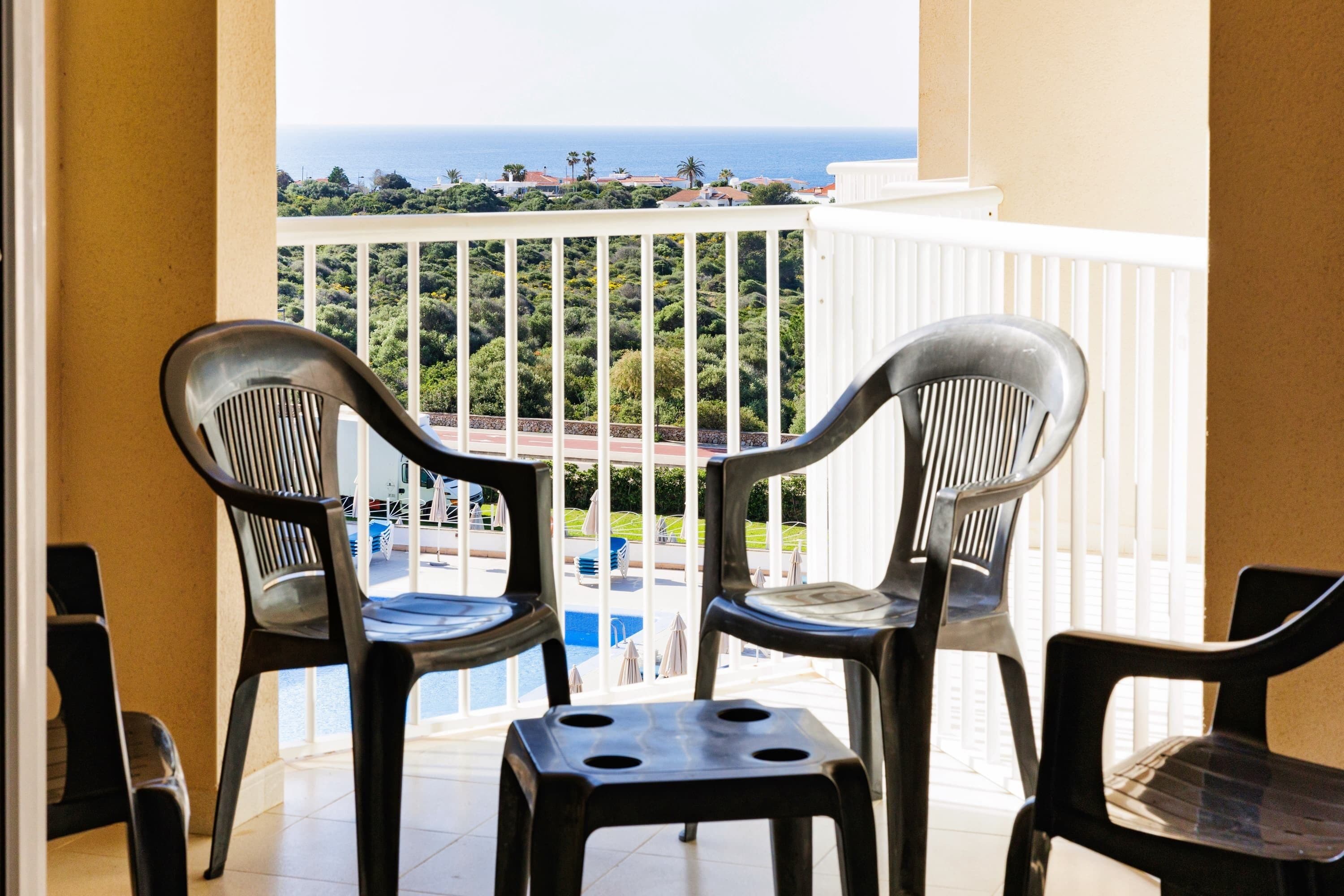 Un balcón con dos sillas de plástico y una mesita pequeña, con vista a una piscina y follaje.