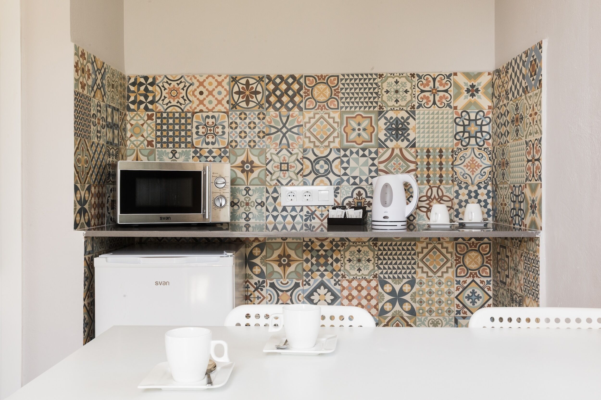 A kitchenette with a microwave, kettle, and coffee maker on a shelf, a white refrigerator below, and a table with a cup and saucer.