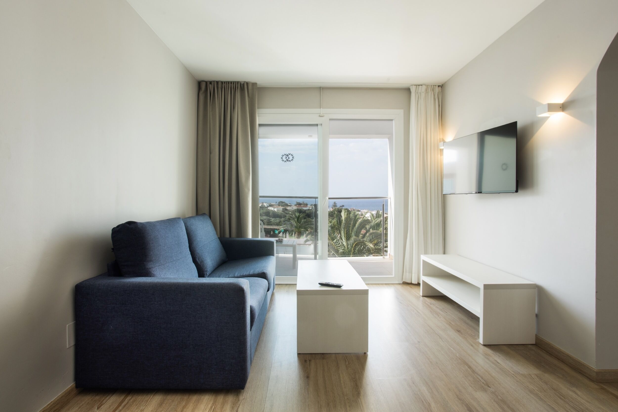 A modern living room with a sofa, a coffee table, a TV mounted on the wall, and a balcony with a view.