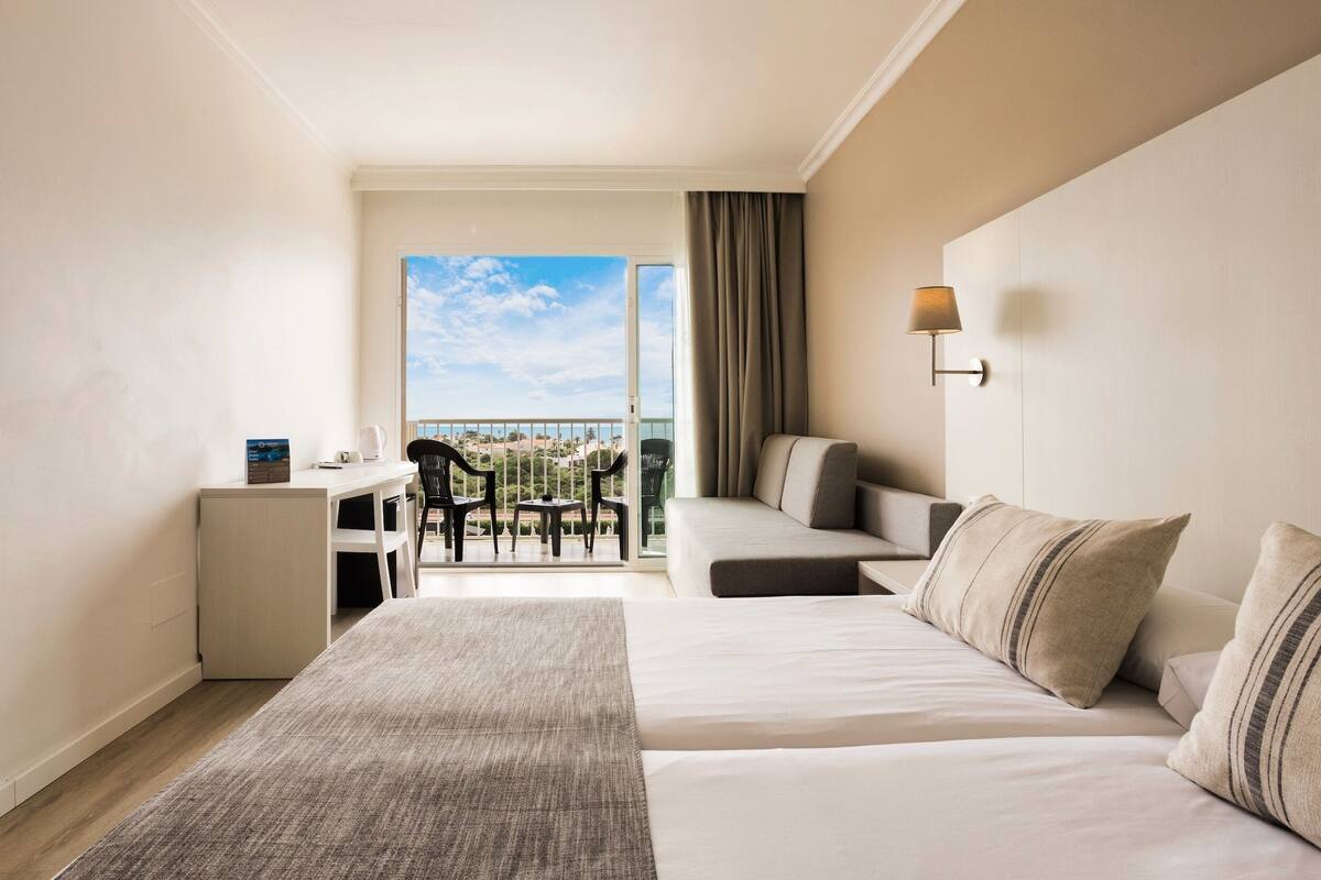 A hotel room with a bed, a desk, a chair, a sofa, and a balcony with a view.