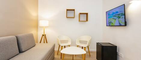 A modern living room with a grey sofa, two white chairs, a small black table, a floor lamp, two picture frames, and a TV mounted on the wall.