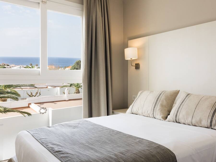 Habitación de hotel con cama, ventana con vista al océano, lámpara de pared y cortina.