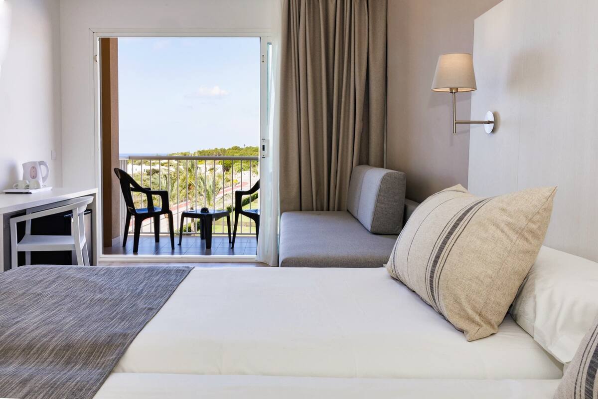 A hotel room with a bed, a desk, a chair, a balcony with a view, and a wall-mounted lamp.