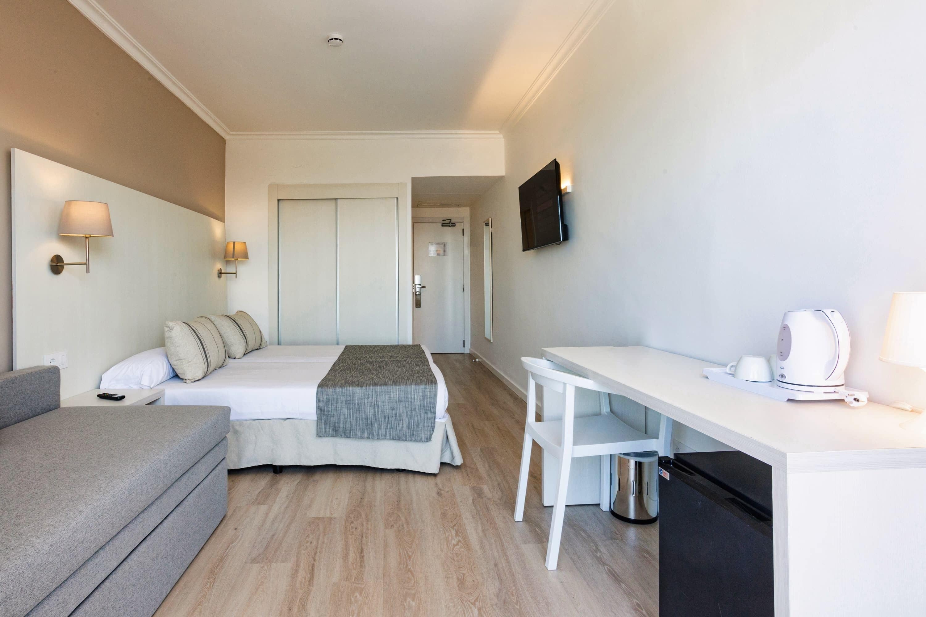 A hotel room with a bed, a sofa, a desk with a kettle, and a TV on the wall.